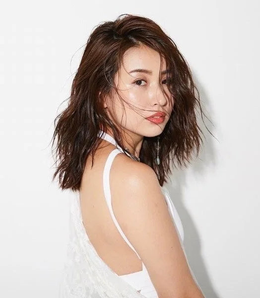 白い背景の前で、肩を出し白いトップスを着た女性のポートレート。濡れたようなウェーブのショートヘアが特徴で、カメラに視線を送っています。自然体で洗練された雰囲気が漂います。