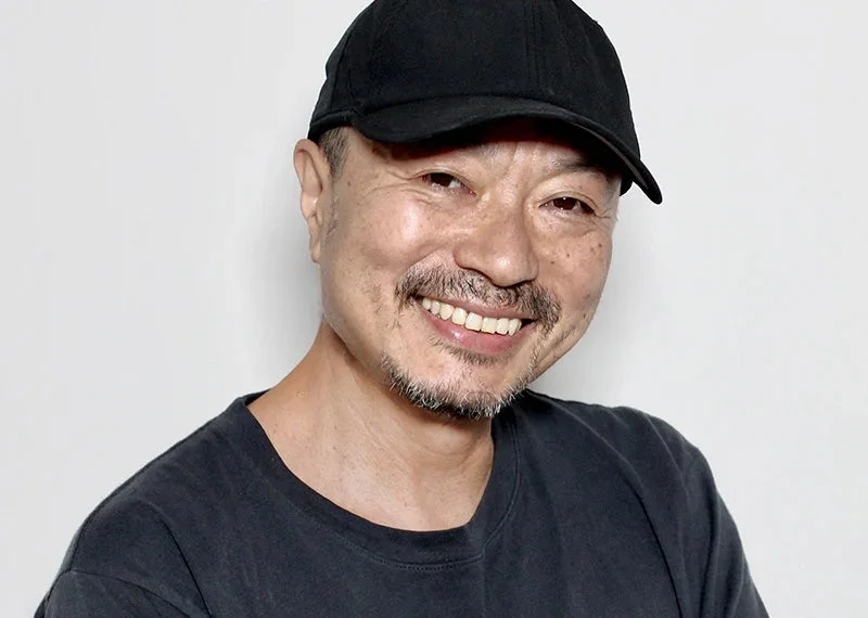 黒いキャップとTシャツを着用した中高年の日本人男性が、白い背景の前で満面の笑みを浮かべているポートレート写真です。ひげと口ひげも特徴的です。