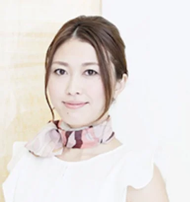 明るい背景の前で微笑むアジア人女性のポートレートです。白いトップスを着用し、首元にはチェック柄のスカーフを巻いています。