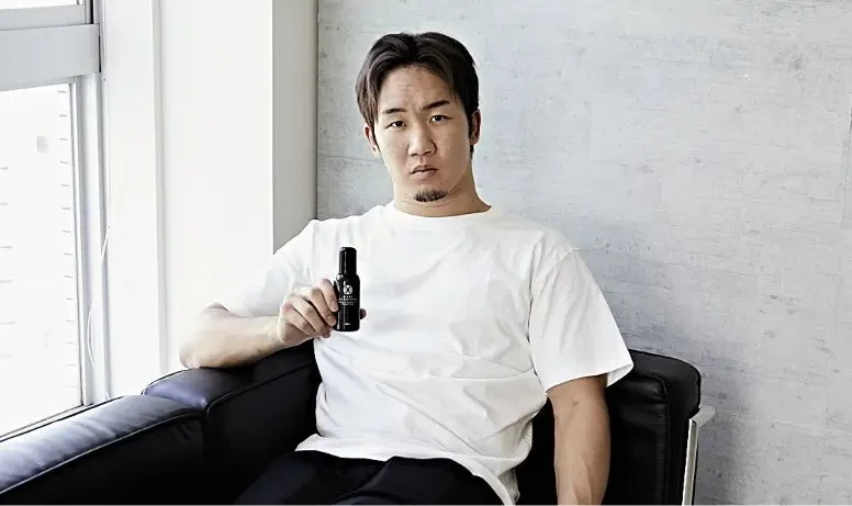 白いTシャツを着た男性が黒いソファーに座り、カメラに向かって黒い育毛剤のボトルを見せています。窓からの光が差し込む明るい室内での一枚です。