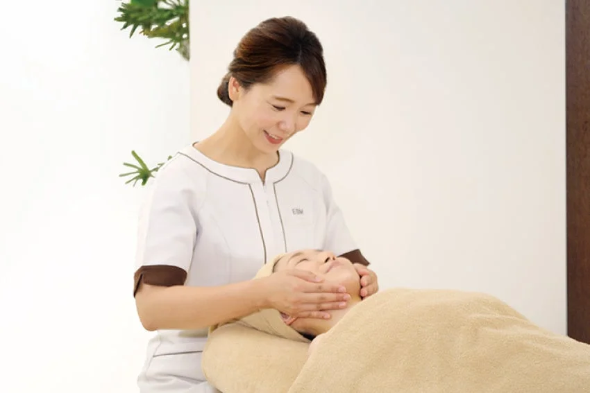 白い制服を着たエステティシャンが、施術台に横たわる女性の顔を優しくマッサージしている様子