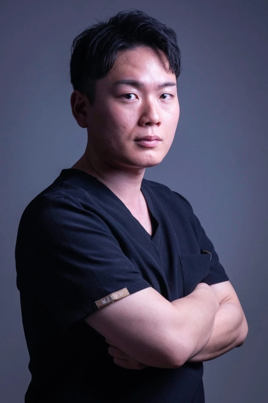 黒いスクラブを着用したアジア人男性のポートレート。腕を組み、真剣な表情でカメラを見つめています。プロフェッショナルな雰囲気が感じられます。