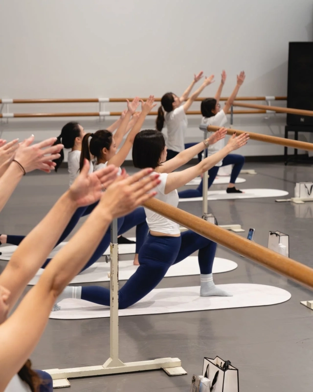 明るいスタジオで、複数の女性がバレエバーを使ってフィットネスを行っている様子