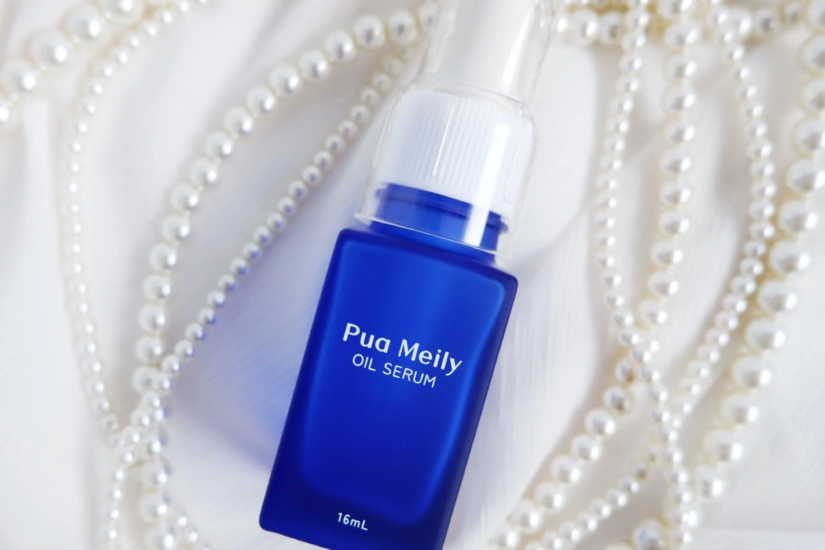 青いボトルに入ったPua Meilyのオイルセラムが、白い布の上に置かれた真珠のネックレスに囲まれています。